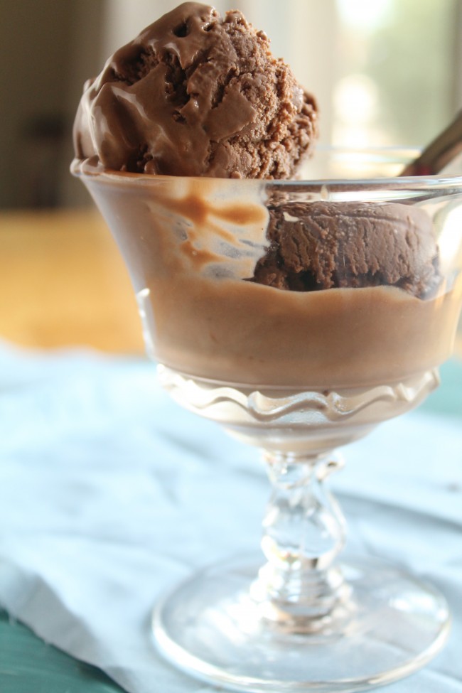 Spoon taking a bite of chocolate ice cream out of a glass serving dish as it begins to melt.