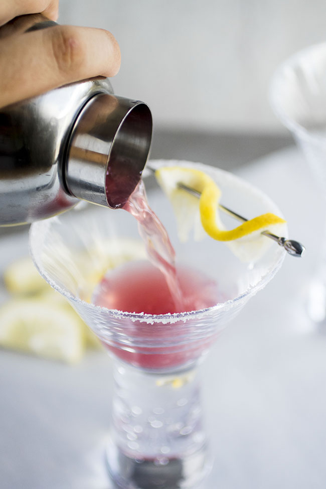 Metal cocktail shaker pouring pink liquid into a martini glass.