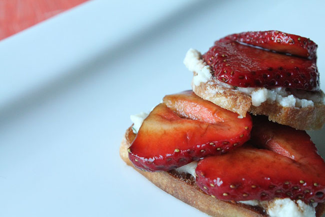 Two baguette slices topped with ricotta cheese and strawberry slices on a white plate.