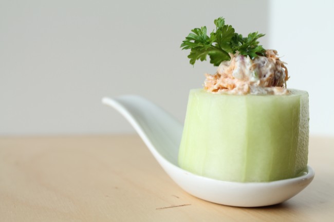 Small white appetizer spoon holding a cucumber cup stuffed with smoked salmon dip and topped with fresh parsley.