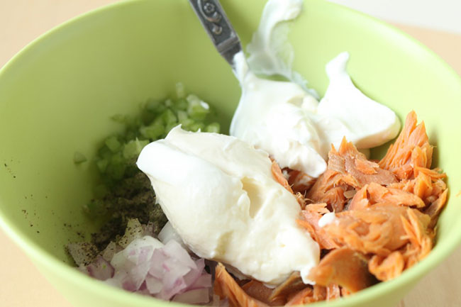 Spoon stirring mayonnaise, smoked salmon, onion, and celery together in a small green bowl.