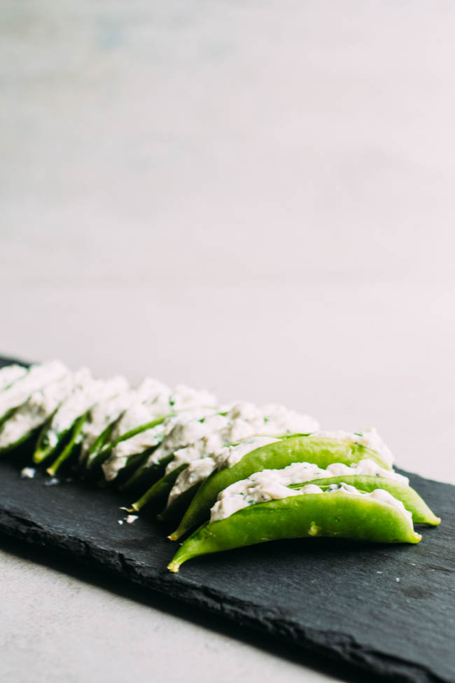 Snap peas stuffed with a cheese and herb mixture arranged on a slate serving platter in front of a white wall.