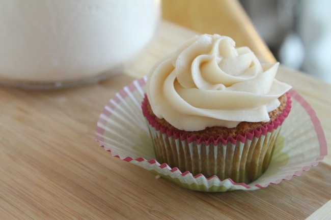 Vanilla cupcake with white frosting on a wooden cutting board next to a jar of powdered sugar.