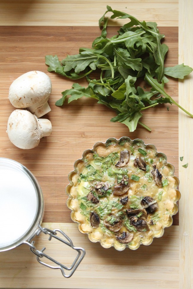 Small mushroom quiche sitting on a wooden cutting board next to a pile of fresh arugula and several whole mushrooms.