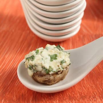 White appetizer serving spoon holding a mushroom stuffed with cheese and herbs.