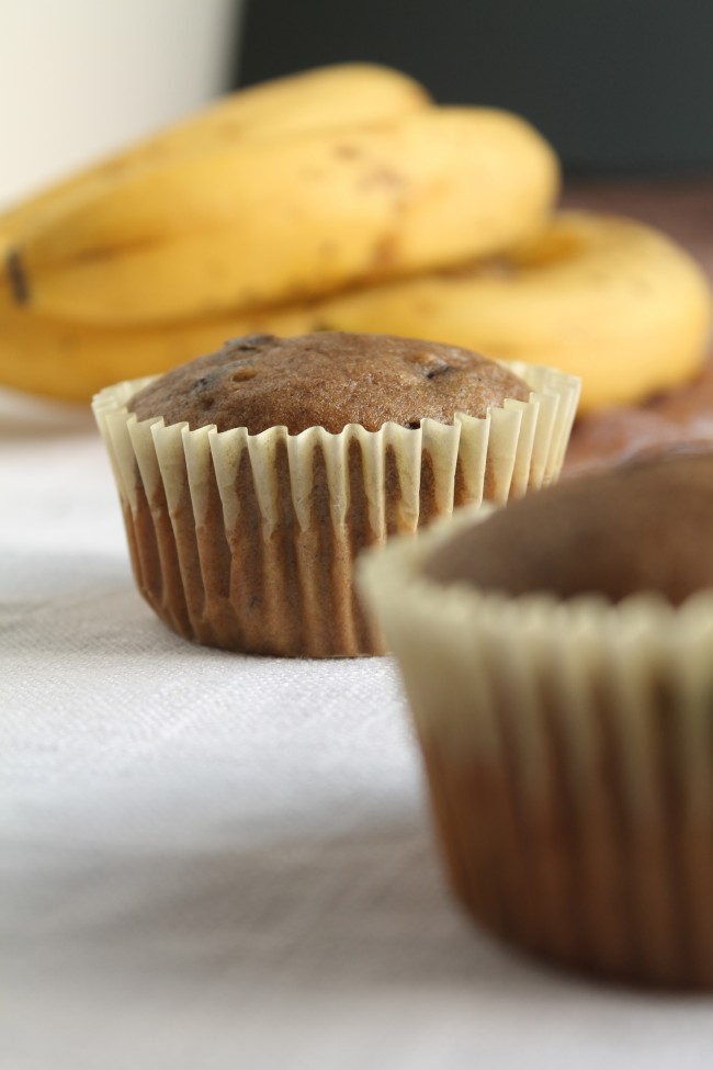 Dark brown muffin in a light yellow wrapper next to a bunch of bananas.