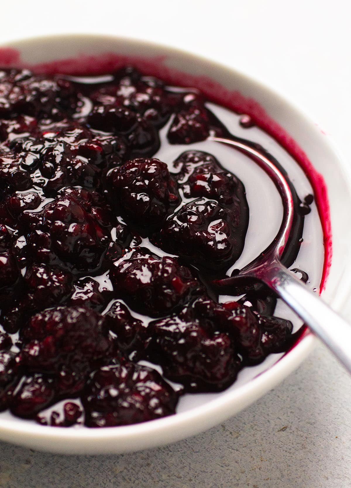 Spoon stirring blackberry sauce in a white bowl.