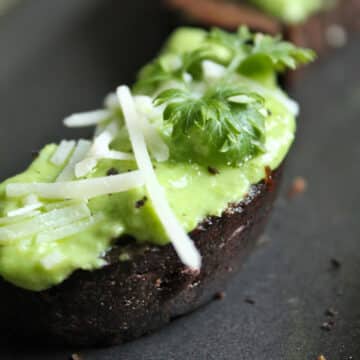 Close up of a piece of pumpernickel toast topped with bright green dip and a sprig of fresh parsley.