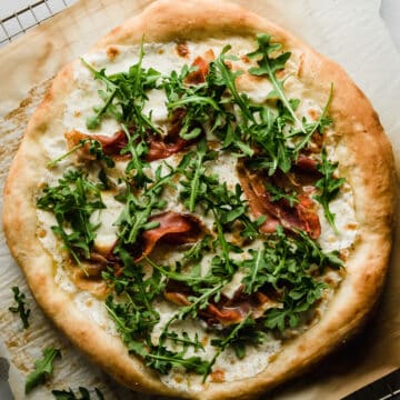 White pizza topped with prosciutto, arugula, and mozzarella, sitting on a wire cooling rack.
