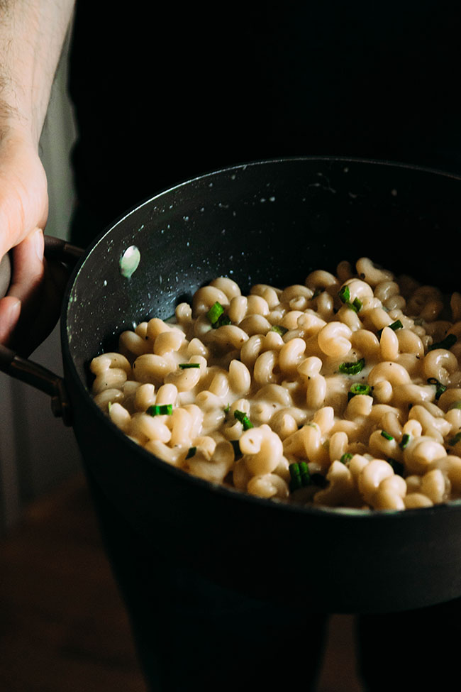 Man\'s hands holding a pot full of macaroni and cheese with green onions on top.