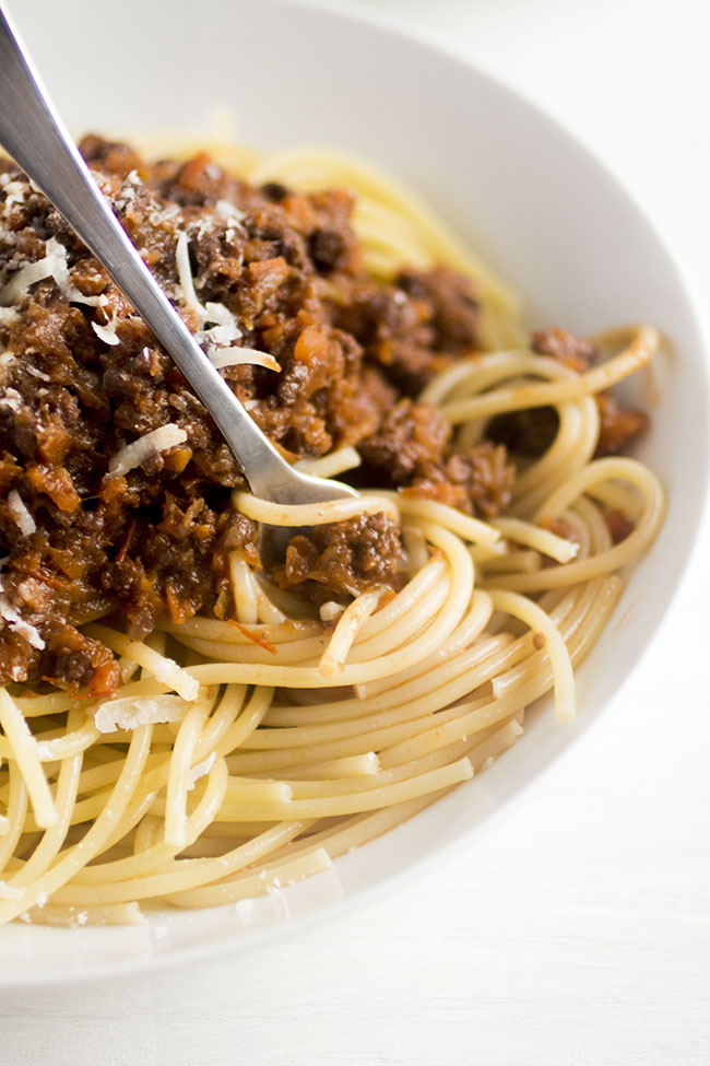 Silver fork twirling spaghetti strands with dark red bolognese sauce in a white bowl.