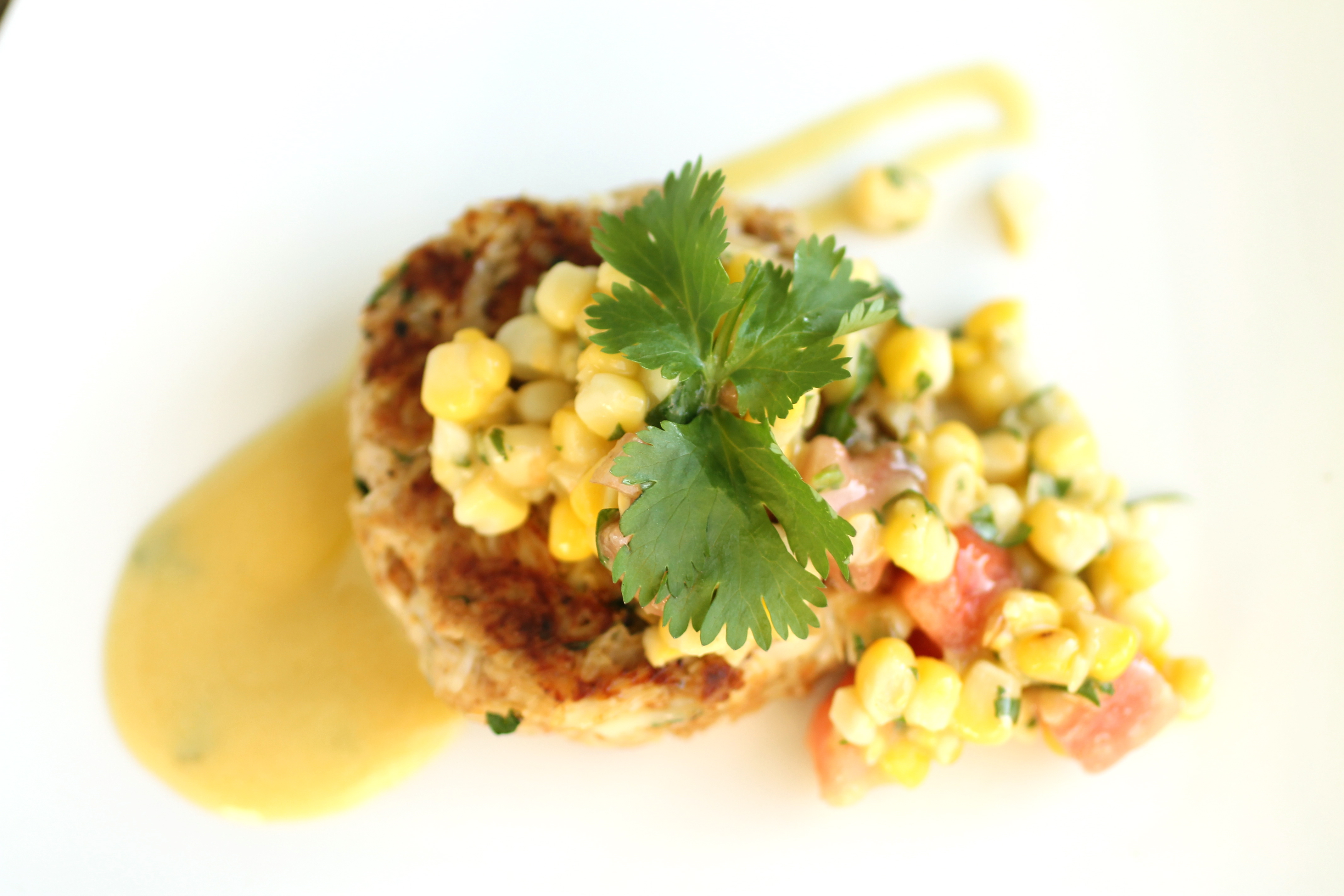 Crab cake topped with corn salsa and a sprig of fresh cilantro atop a white plate with a dollop of mustard sauce.