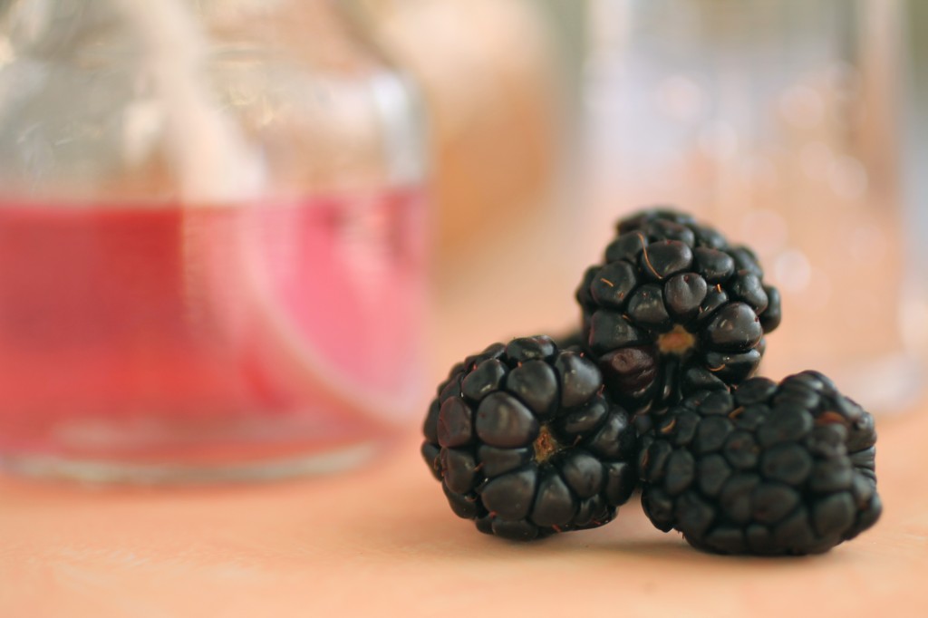 Fresh blackberries on a peach colored surface next to a small glass jar filled with blackberry vodka.