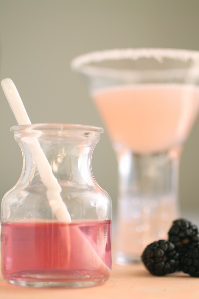 Small jar of blackberry infused vodka in front of a martini glass filled with a light pink cocktail.
