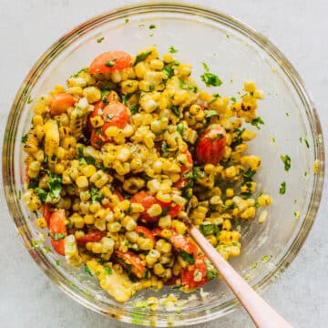 Copper spoon stirring grilled corn, tomatoes, and cilantro together in a glass bowl.