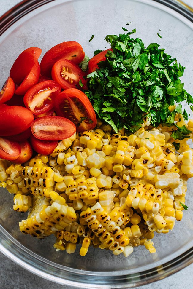 Halved cherry tomatoes, chopped cilantro, and grilled corn in a glass bowl.