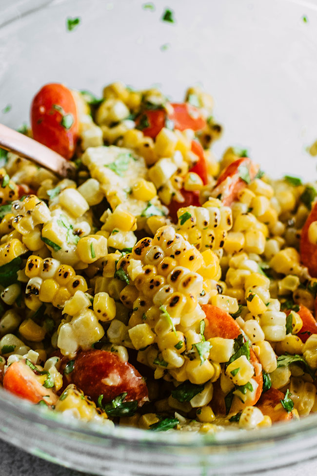 Rose gold spoon stirring grilled corn and tomatoes together in a glass bowl.