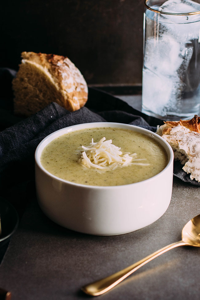 Broccoli soup in a white bowl, in front of a loaf of crusty bread and a blue napkin.