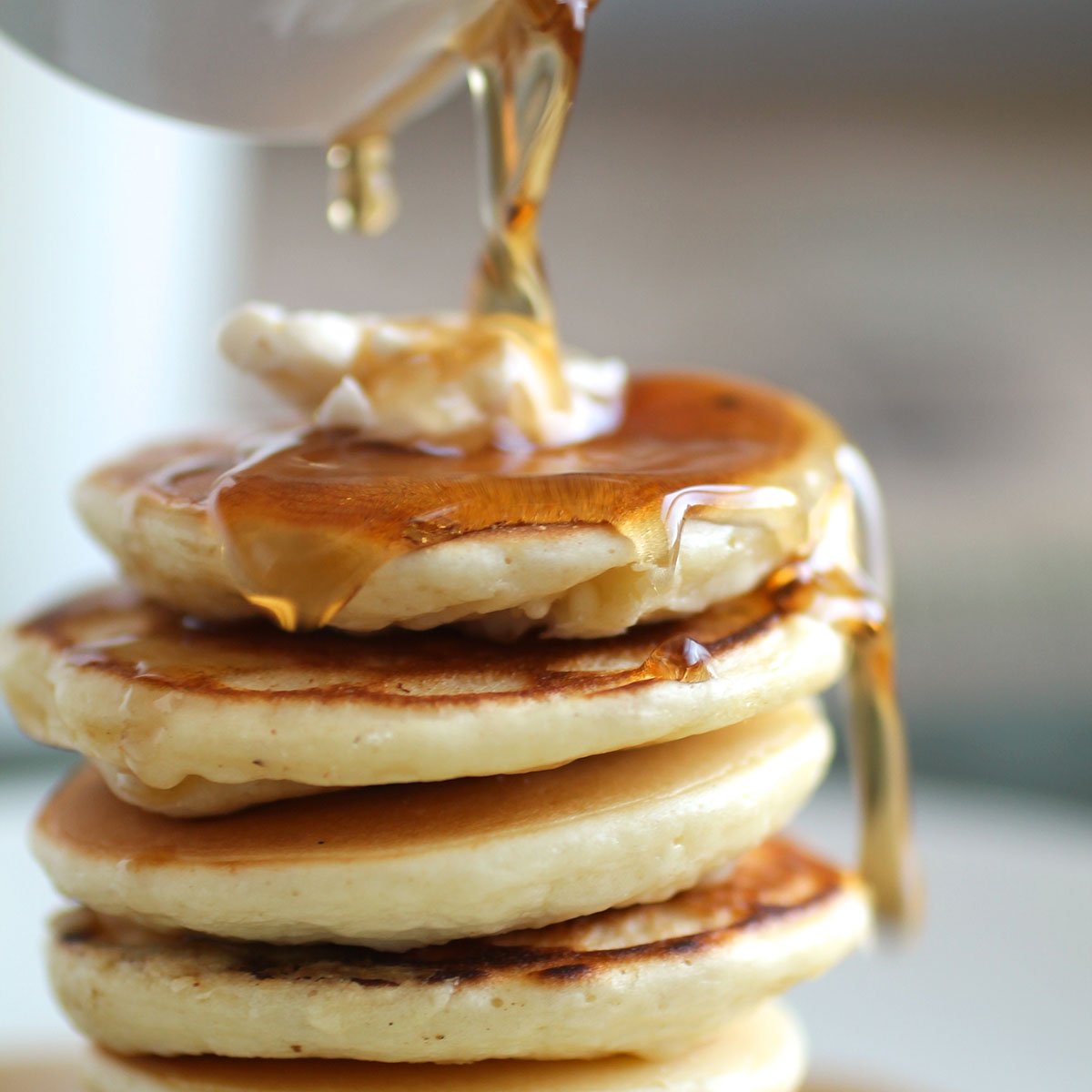 White dish pouring maple syrup over a stack of pancakes.