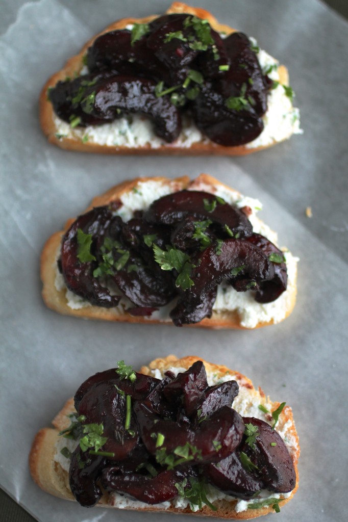 Close up of a crostini topped with cheese spread, roasted mushrooms, and fresh parsley on a light surface.