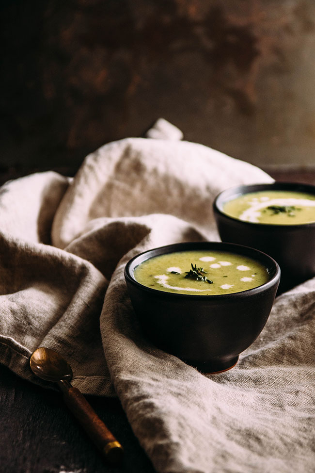 Green soup in a small black bowl, next to a a light brown linen napkin.