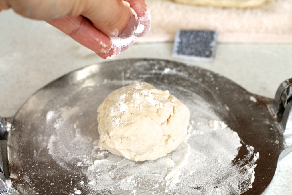Hand sprinkling flour over tortilla dough that\'s sitting on a tortilla press.
