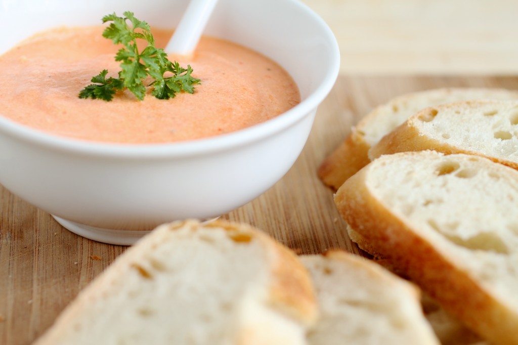 Close up of a white bowl filled with red pepper dip sitting next to a pile of baguette slices.
