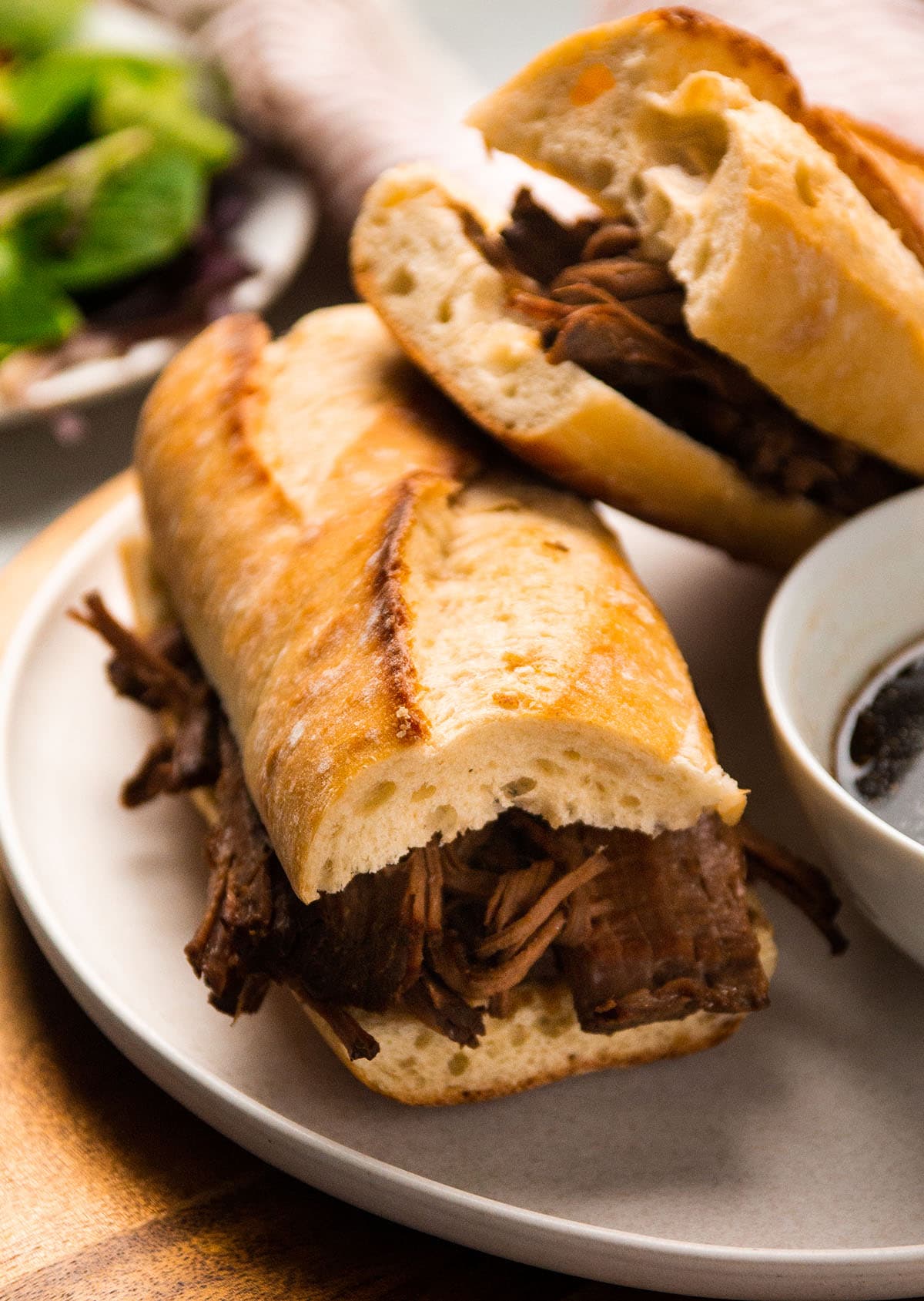 Two french dip sandwiches on a white plate next to a bowl of au jus.