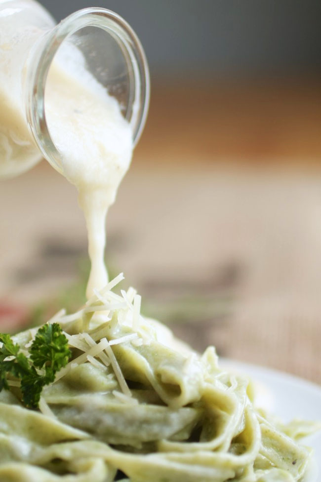 Glass jar pouring alfredo sauce over spinach fettuccine.
