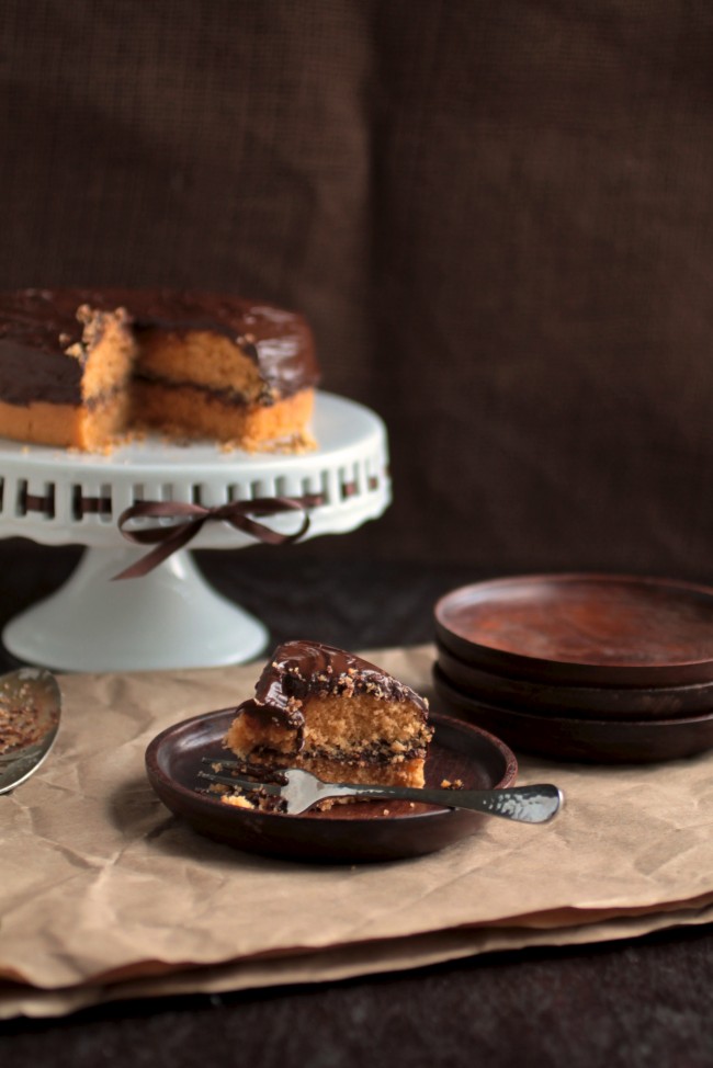 Slice of cake on a wooden plate in front of a white cake stand.