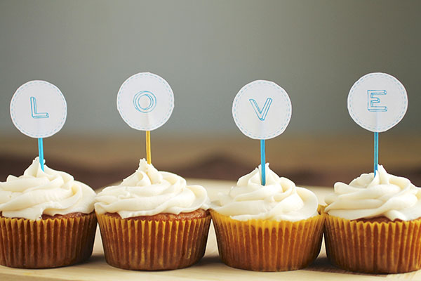 Four vanilla cupcakes, each holding a toothpick with a letter decoration to spell \'love\'.