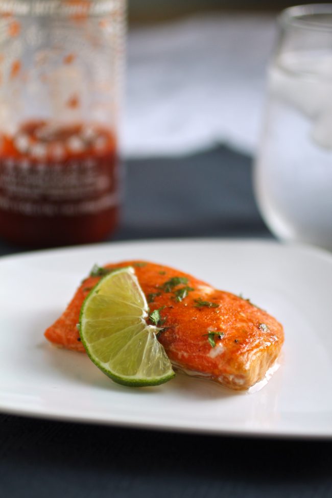 Slice of salmon on a white plate next to a lime wedge, in front of a bottle of sriracha.