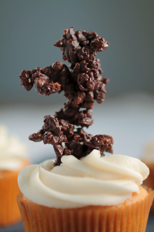 Vanilla cupcake with a chocolate rice krispy branch stuck into the frosting.