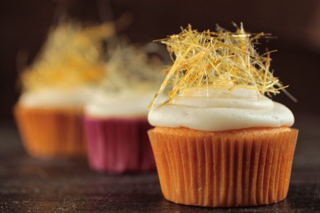 Three cupcakes with vanilla frosting, topped with a small nest of thin sugar strands.