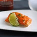 Slice of salmon on a white plate next to a lime wedge.