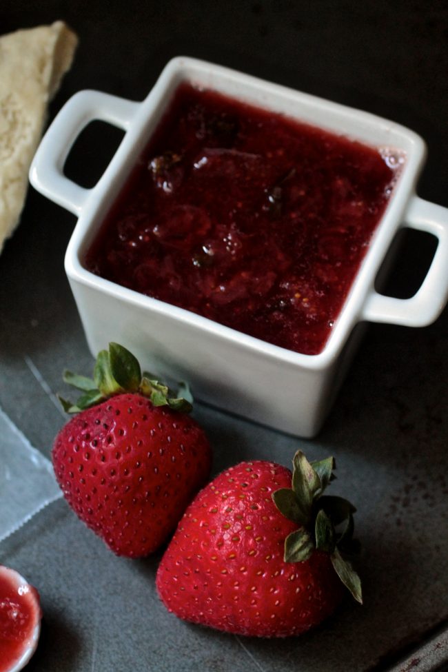 Strawberry jam in a small white dish, next to several fresh strawberries.