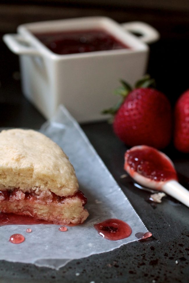 A scone with strawberry jam next to fresh strawberries and a spoon filled with jam.
