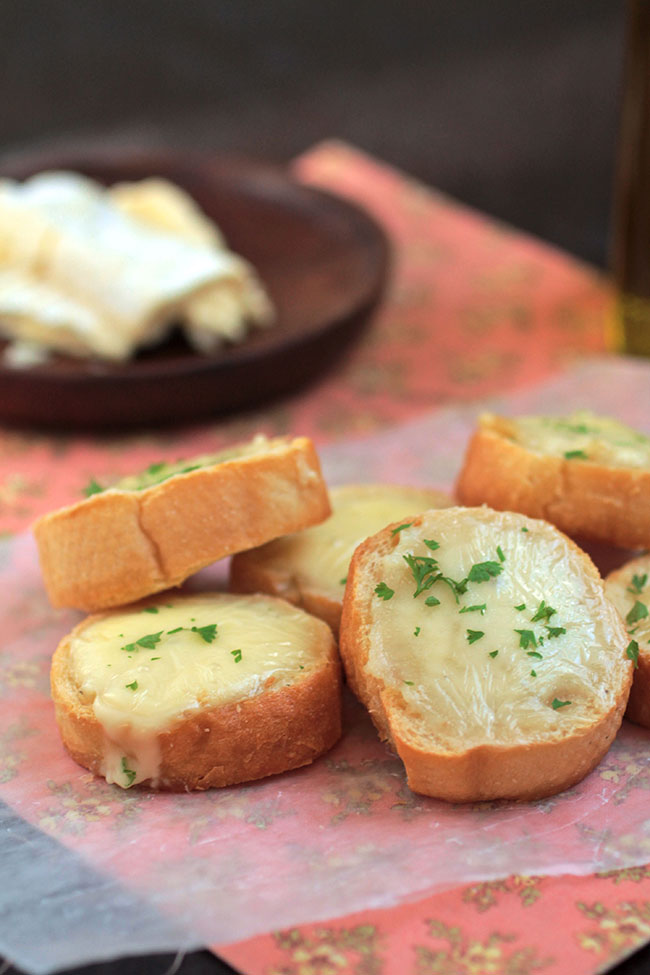 A pile of crostini sitting on a pink napkin, next to a wooden plate with a piece of brie on it.