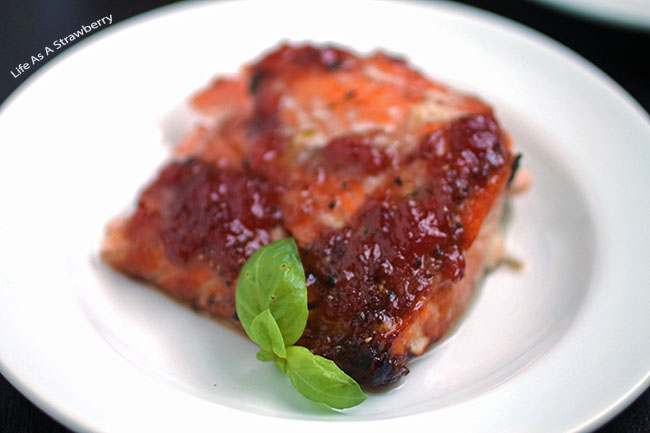 Slice of glazed salmon on a white plate, with a sprig of fresh basil on top.