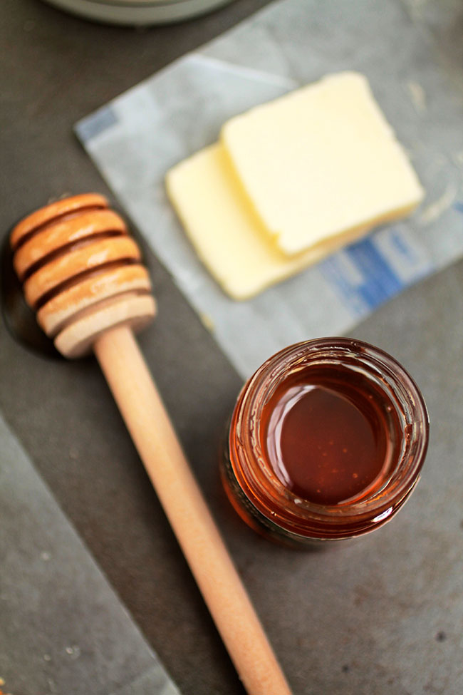 A jar of honey next to several pats of butter and a honey dipper.