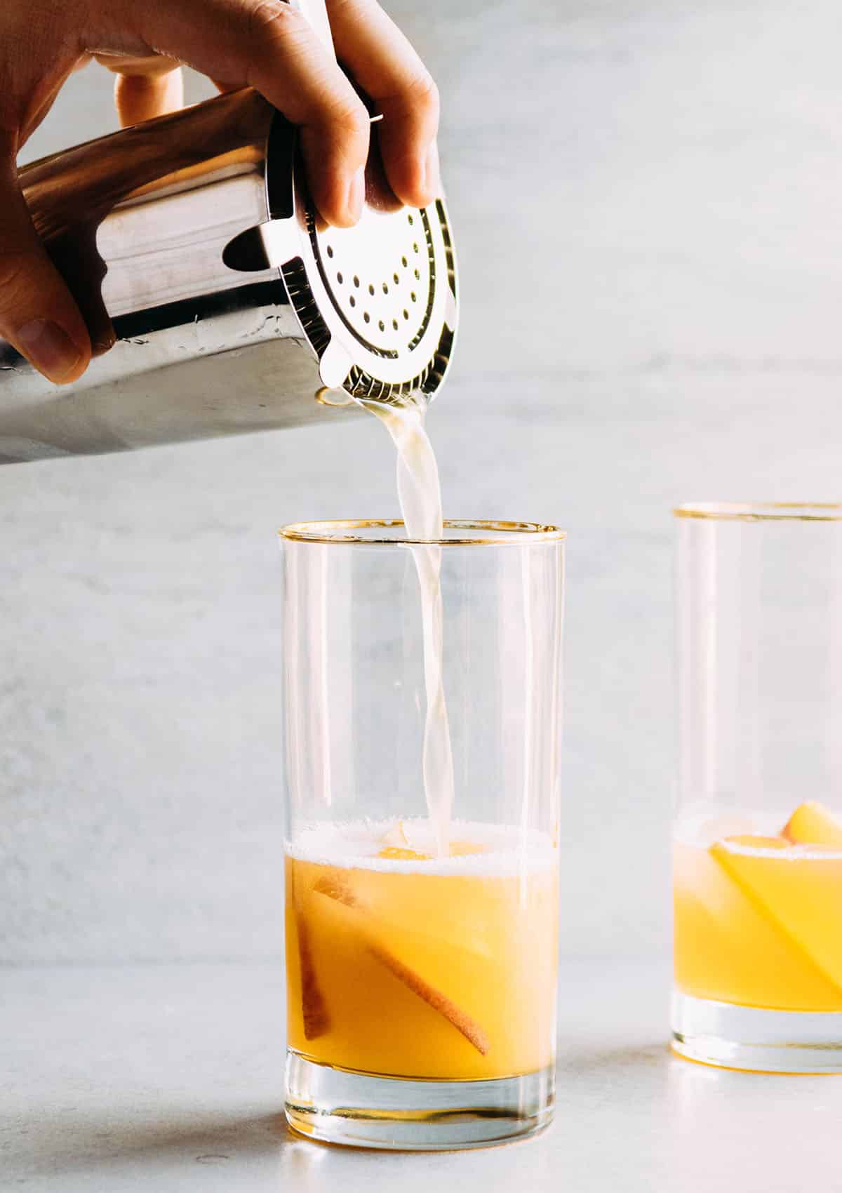 Hand pouring peach cocktail into a tall glass from a metal cocktail shaker.