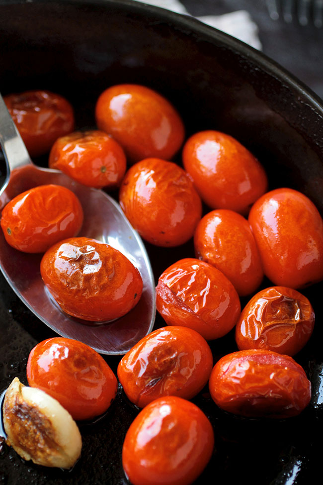 Silver spoon stirring blistered tomatoes in a black skillet.