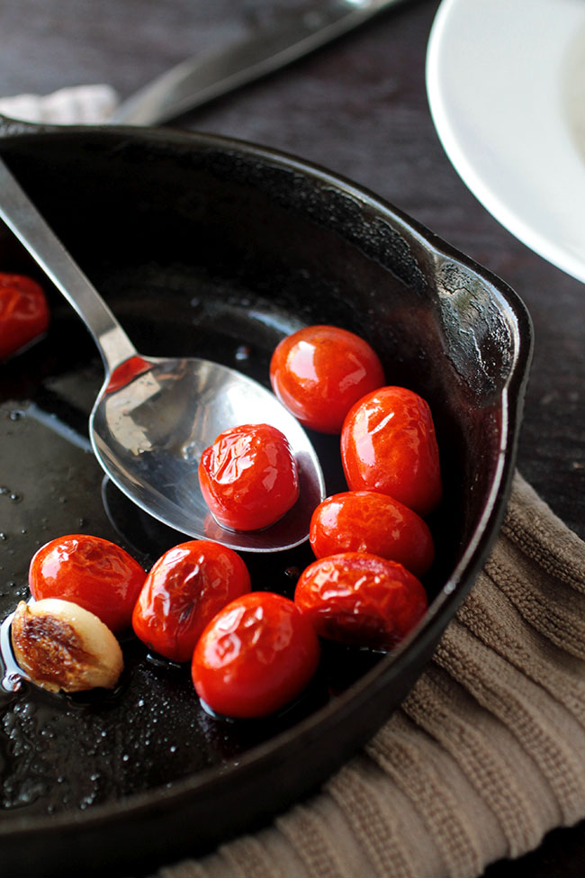 Spoon stirring blistered tomatoes in a cast iron skillet.