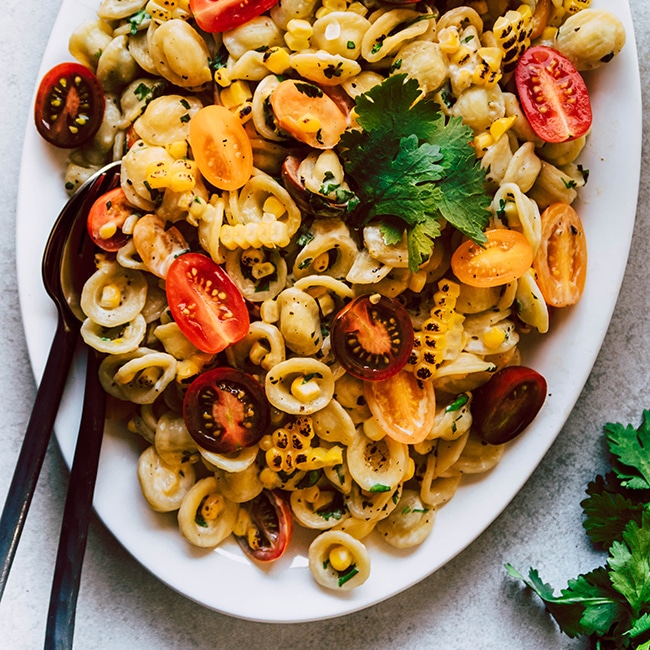 Orecchiette pasta with corn and tomatoes on a white oval platter with black serving utensils.