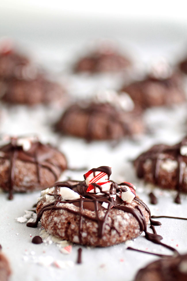 Chocolate cookies topped with ganache, crushed peppermint, and chocolate drizzle, on a white surface.