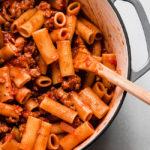 Wooden spoon stirring rigatoni and tomato sauce in a light grey pot.