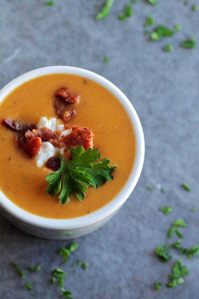 Butternut squash soup in a white bowl, topped with bacon, crumbled goat cheese, and fresh parsley.