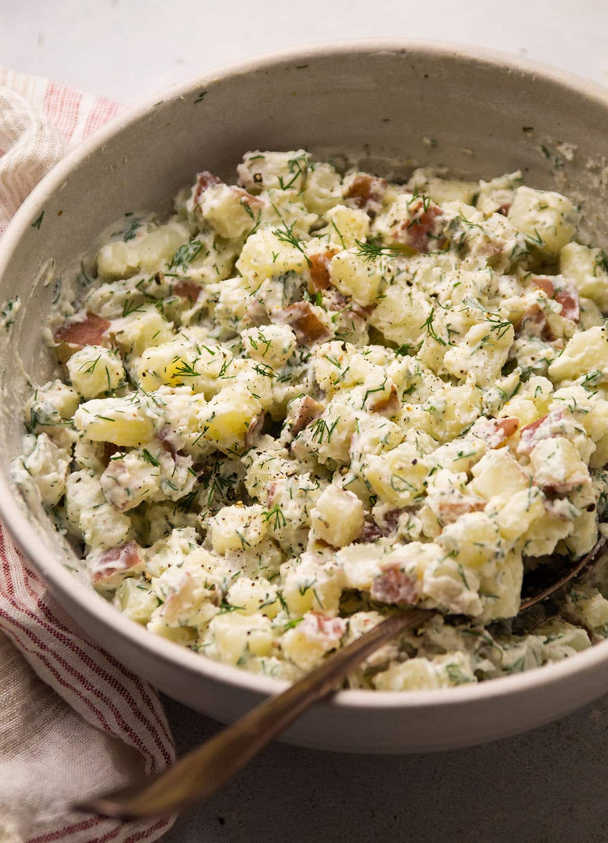 Copper spoon lifting potato salad out of a white bowl.