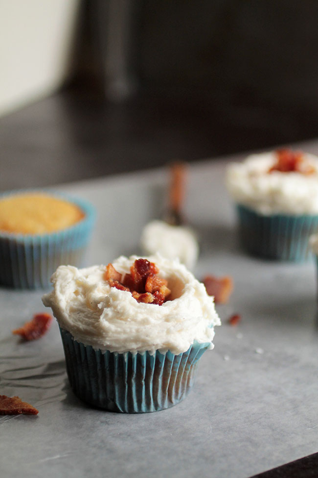 Cupcake in a blue wrapper topped with fluffy frosting and crumbled bacon.