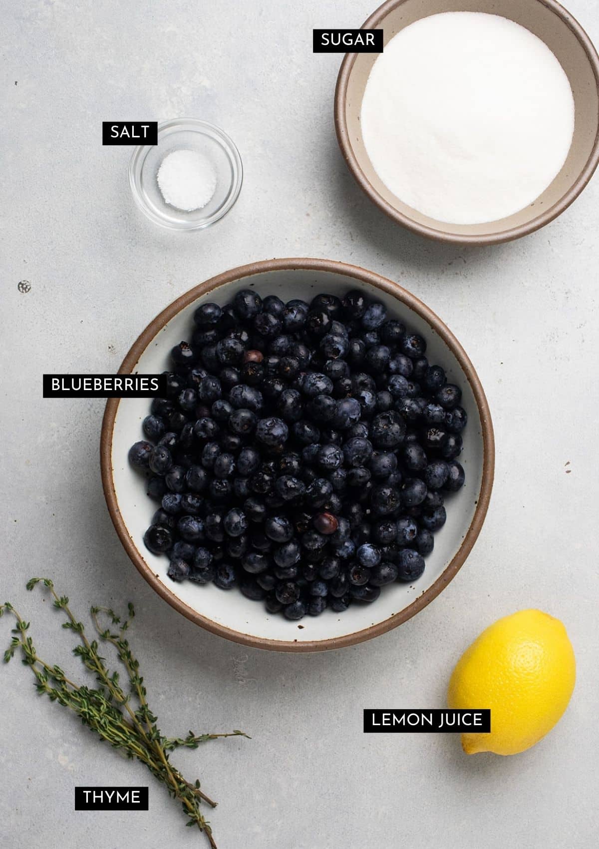Blueberries, thyme, sugar, and a lemon on a white table.